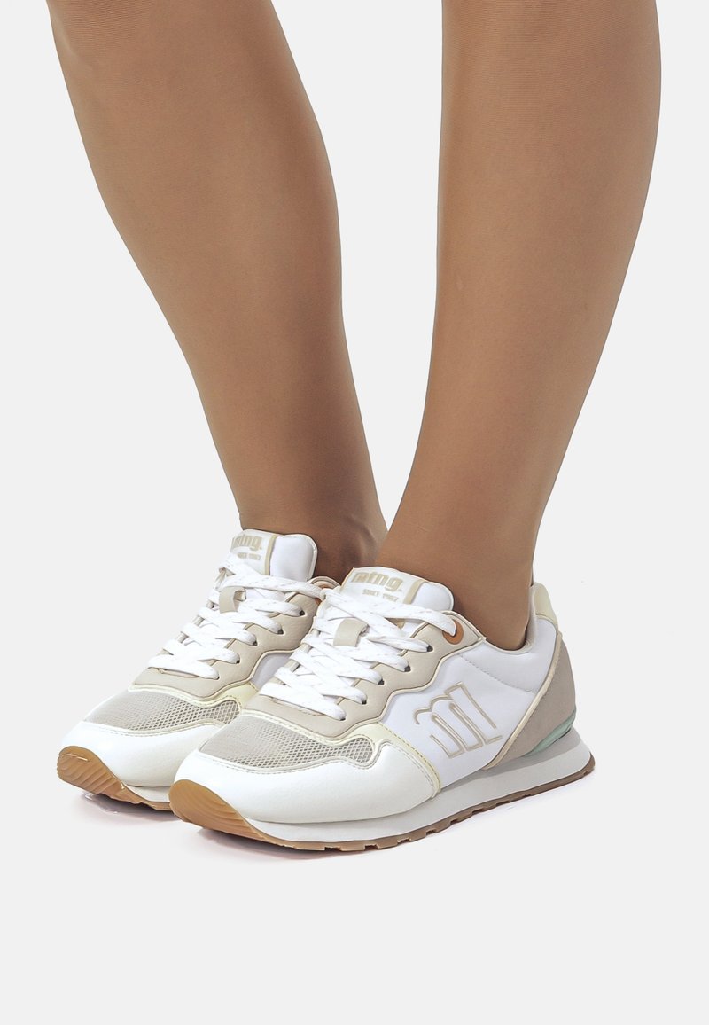 Zapatillas deportivas blancas con acentos beige, paneles de malla y suela de goma. Presentan un diseño de cordones y una marca en el lateral y la lengüeta.