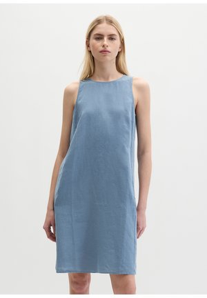 Frau steht und trägt ein ärmelloses, knielanges, hellblaues Kleid vor einem einfarbigen weißen Hintergrund.