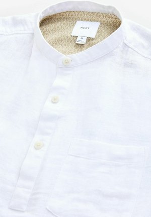 Camisa de lino blanca con cuello mao, que presenta un bolsillo delantero y botones claros. El interior cuenta con un forro beige con patrón.