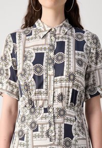 Femme portant une robe chemise à manches courtes avec un motif géométrique composé de cercles et de carrés en noir, blanc, beige et vert atténué.