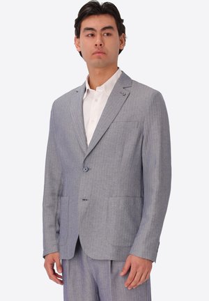 Uomo che indossa una giacca da completo grigio chiaro e pantaloni abbinati sopra una camicia bianca con colletto, rivolto leggermente a sinistra con espressione neutra.