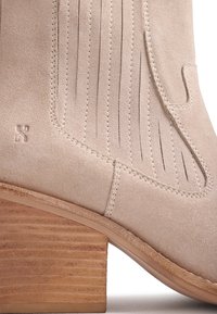 Κοντινή λήψη μποτάκι αστραγάλου από μπεζ σουέτ με στιβαρό ξύλινο τακούνι και διακοσμητικά σχέδια με ραφές και περικοπές στο πλάι.