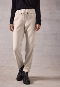 Beige, elastische broek met tailleband en trekkoord, zijzakken en toelopend broek ontwerp. Draagt met zwarte haklaarzen.