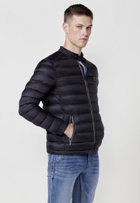 Chaqueta de plumas negra con cuello alto, diseño de acolchado horizontal y dos bolsillos laterales con cremallera, llevada sobre unos vaqueros azules.