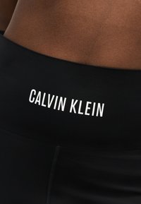 Μαύρο ένδυμα με ψηλή μέση και λευκό λογότυπο "Calvin Klein" στη μέση, φορεμένο από άτομο με καφετί επιδερμίδα.