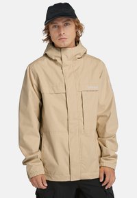 Beige vattentät jacka med dragkedja i fronten, hög krage och elastiska ärmslut. Har Timberland-logotyp och två bröstfickor. Svart keps på.