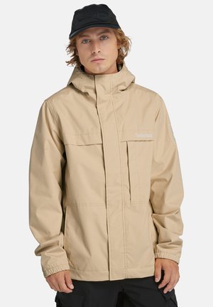 Giacca impermeabile beige con zip frontale, colletto alto e polsini elasticizzati. Presenta il logo Timberland e due tasche sul petto. Cappello nero indossato.