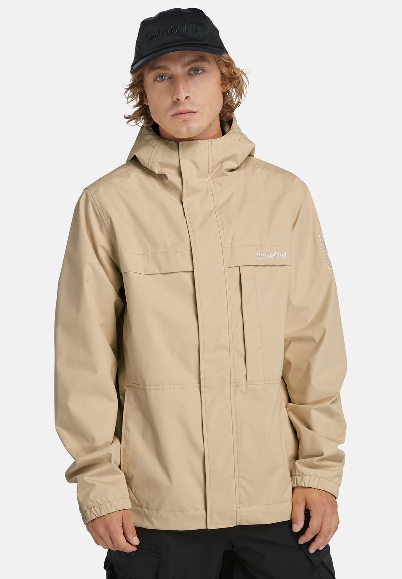 Beige vattentät jacka med dragkedja i fronten, hög krage och elastiska ärmslut. Har Timberland-logotyp och två bröstfickor. Svart keps på.