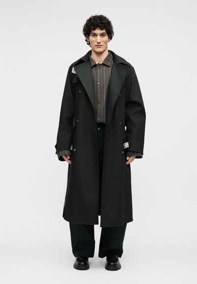 Άνδρας με σγουρά μαλλιά φορώντας ένα μακρύ μαύρο διπλό σακάκι πάνω από ένα καφέ ριγέ πουκάμισο με κουμπιά, μαύρο παντελόνι με φαρδιά ποδάρια και μαύρα παπούτσια.