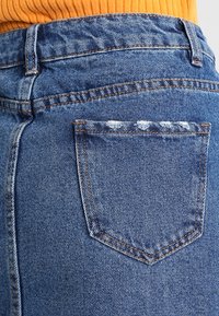 Spijkerbroek met hoge taille in een vervaagde blauwe wassing, voorzien van een achterzak met rafelige details en contrasterende oranje stiksels.