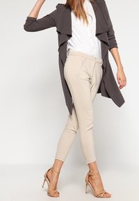 Lång, öppen grå kavaj över en blus med vita prickar, tillsammans med krämfärgade tapered-byxor och nätta sandaler med hög klack i nude.