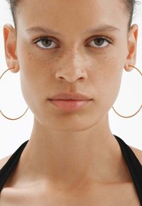 Aros de oro, grandes y circulares, llevados con una blusa halter negra. La piel tiene una textura suave con pecas visibles. Fondo neutro.