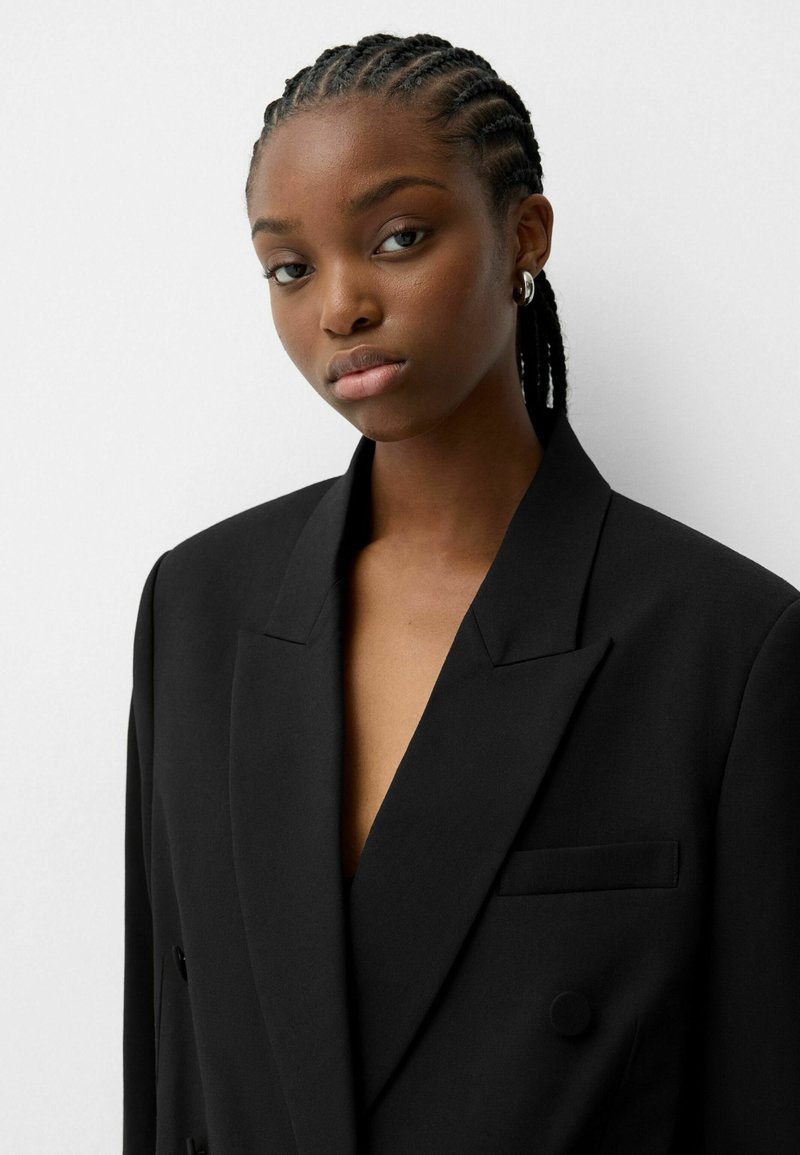 Blazer noir avec revers crantés, design à double boutonnage, tissu texturé et une poche poitrine. Lignes épurées et coupe ajustée.