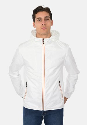 Uomo che indossa una giacca bianca con cappuccio, con cerniere frontali e delle tasche bordate di arancione, in piedi con le mani dietro la schiena su uno sfondo semplice.