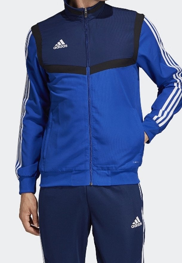 Blå atletisk jacka med dragkedja, med marinblå överpanel och svarta detaljer. Har vitt Adidas-logotyp och tre ränder på ärmarna.