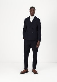 Giacca da abito doppiopetto blu navy con rever a pattina, pantaloni coordinati su misura e scarpe in pelle marrone. Camicia bianca sotto.