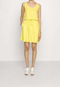 Gul ärmlös klänning med dragsko i midjan, avslappnad passform och sidofickor. Bärs med beige sneakers mot en neutral bakgrund.