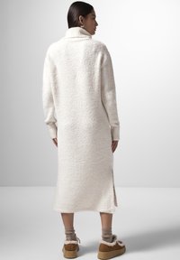 Abito maglione lungo, color crema, con texture, alto colletto e spacchi laterali, abbinato a scarpe slip-on marroni e calzini alla caviglia.