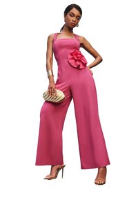 Fuchsia jumpsuit met wijde pijpen, vierkante halslijn en een grote bloemaccent bij de taille. Model met een gouden clutch in de hand, draagt metallic hakken.