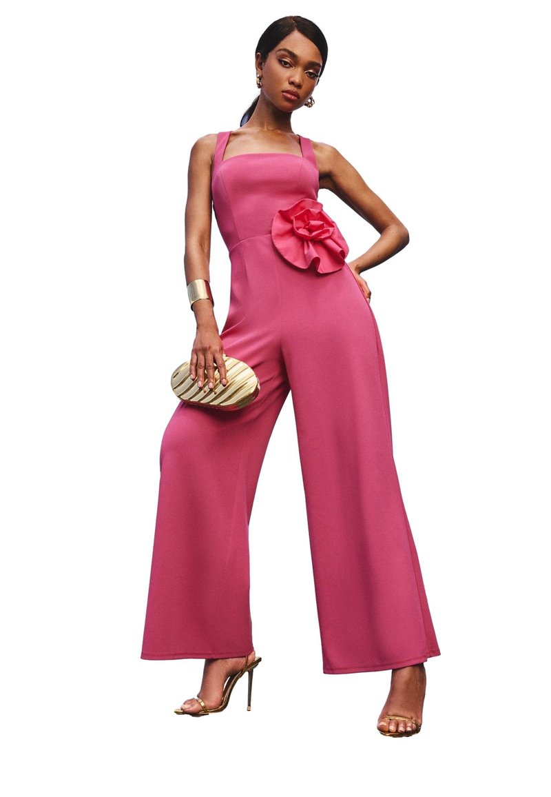 Fuchsia jumpsuit met wijde pijpen, vierkante halslijn en een grote bloemaccent bij de taille. Model met een gouden clutch in de hand, draagt metallic hakken.