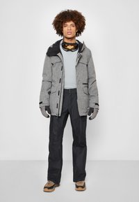 Grijze gestreepte winterjas met zwarte accenten, gecombineerd met donkere broeken en handschoenen. Bruine wandelschoenen maken de outfit af.