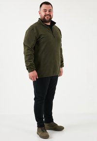 Fleece pullover i dyb grøn med en halv-zip krave, parret med sorte bukser og olivengrønne sneakers, med en glat tekstur og elastiske manchetter.