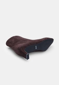 Hnědé semišové kotníkové boty s špičatou špičkou a texturovaným svrškem. Mají černou gumovou podešev a mírný podpatek, což podtrhuje minimalistický design.