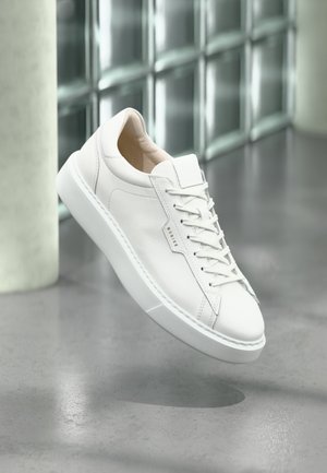 Biały niski sneaker ze sznurówkami i gumową podeszwą, zawieszony nad gładką szarą podłogą w nowoczesnym wnętrzu.