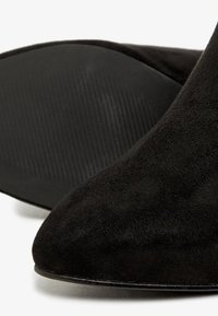 Schwarze Wildleder-Stiefeletten mit spitzen Zehen, einer strukturierten Oberfläche und eleganten schwarzen Gummisohlen. Eine Nahaufnahme hebt die Materialien und die Verarbeitung hervor.