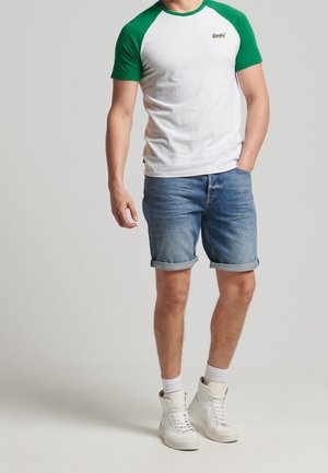 Weißes T-Shirt mit grünen Raglanärmeln, kombiniert mit hochgekrempelten Jeansshorts. Weiße High-Top-Sneaker vervollständigen das Outfit.