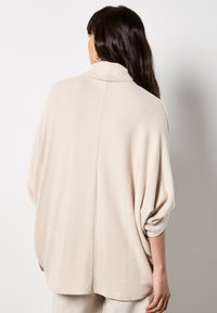 Suéter corto, beige y de gran tamaño con cuello de embudo, hecho de una tela suave. Presenta hombros caídos y un dobladillo redondeado, con una textura lisa.