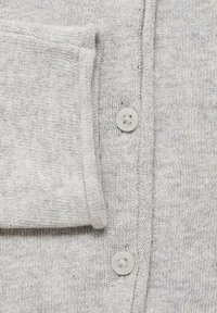 Ljusgrå cardigan i stickad kvalitet med en ficka i patch-stil och två runda, ljusa knappar längs framme på knappslån.