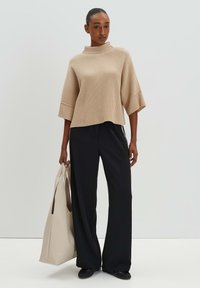 Beiger Rippstrickpullover mit hohem Kragen, kombiniert mit schwarzen, weit geschnittenen Hosen. Das Model hält eine hellbeige Tote-Tasche. Schlichtes, modernes Design.