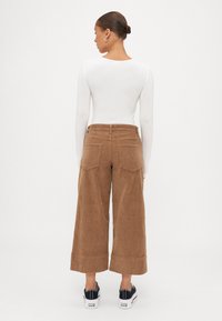 Bruine ribfluwelen broek met wijde pijpen, grote zakken en gestikte manchetten, gecombineerd met zwarte sneakers en een witte top met lange mouwen.