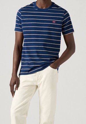 Homme portant un t-shirt à manches courtes rayé bleu marine avec un petit logo rouge, associé à un pantalon beige clair, une main dans la poche.