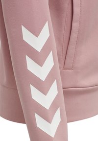 Manche d'un sweat-shirt rose avec quatre bandes blanches en forme de chevrons pointant vers le bas et une poche latérale visible sur le corps.