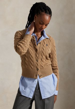 Žena s opletenými vlasmi, ktorá nosí béžový káblíkový sveter cez modrú košeľu na gombíky a sivé nohavice, pózujúca s jednou rukou na krku.