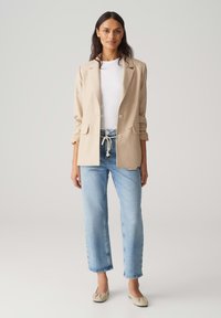 Beige blazer met revers en twee voorzakken, gecombineerd met lichtblauwe ontspannen jeans en beige ballerina's, tegen een neutrale achtergrond.