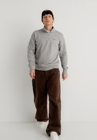 Νέος άνδρας περπατάει, φορά γκρι φούτερ με φερμουάρ στο λαιμό, καφέ παντελόνι φαρδύ, λευκά αθλητικά παπούτσια και μαύρο καπέλο σε απλό λευκό φόντο.