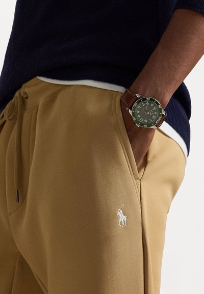 Khaki sweatpants with an embroidered white horse logo, paired with a brown leather strap watch featuring a green dial and silver accents.