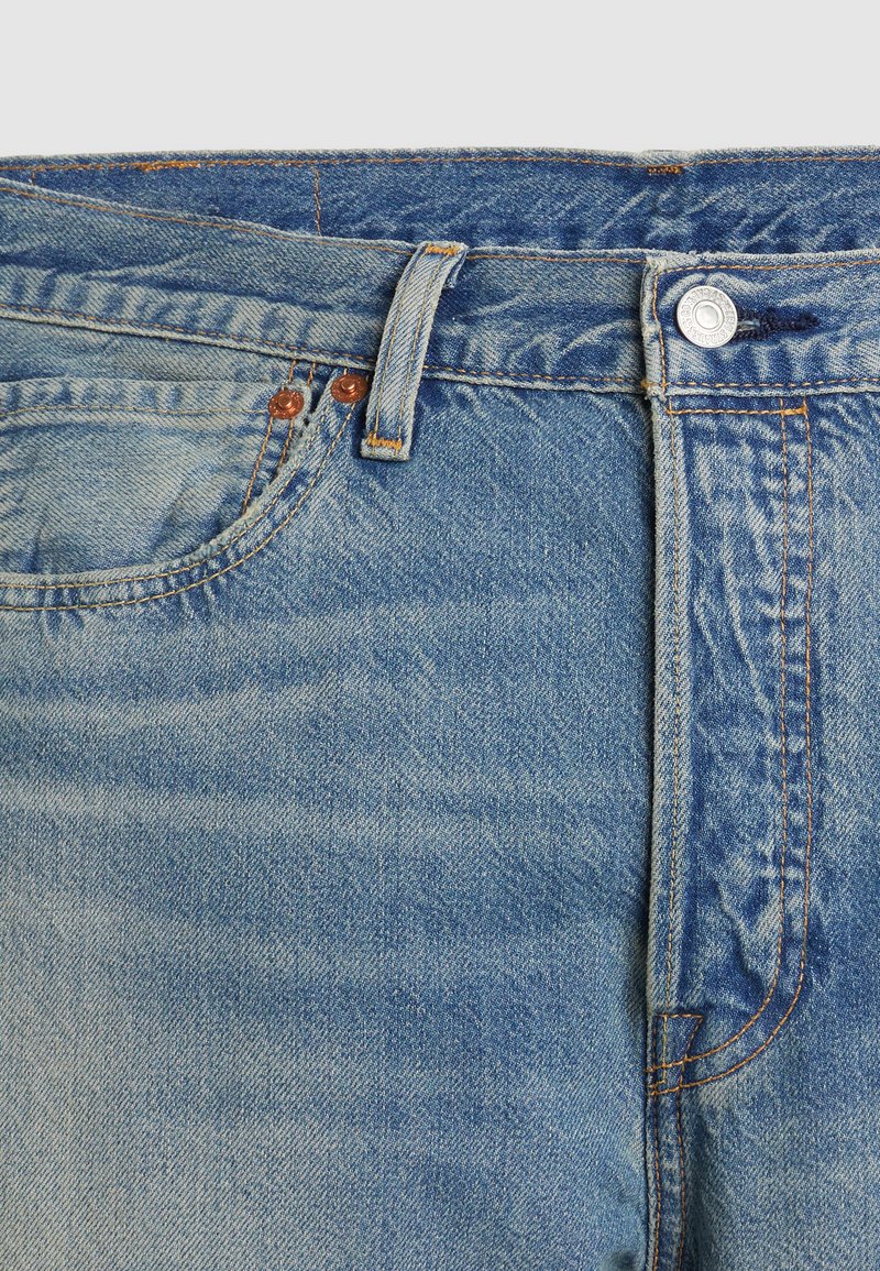 Jean en denim bleu clair avec une texture délavée, doté d'un bouton en cuivre, de surpiqûres orange et de deux poches avant.