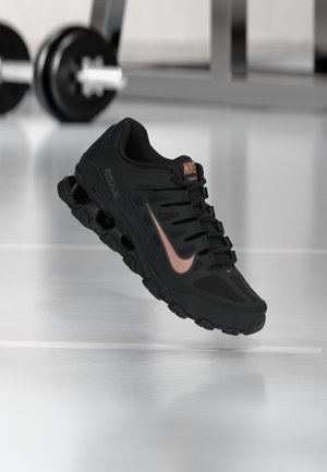 Zwarte Nike trainingsschoen met roségouden swoosh en gedempte zool, zwevend weergegeven tegen een fitnessvloer met gewichten op de achtergrond.