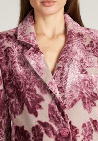 Blazer de terciopelo en taupe claro con un patrón floral en burdeos profundo. Detalles destacados incluyen un acabado texturizado, cuello solapa y un botón en la parte delantera.