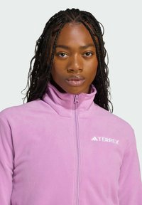 Rosa Fleecejacke mit hohem Kragen und durchgehendem Reißverschluss. Ausgestattet mit einem weißen "TERREX"-Logo auf der linken Seite. Weicher Stoff und enganliegende Passform.