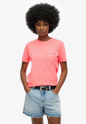 Camiseta rosa coral con cuello redondo y mangas cortas, que presenta un pequeño bolsillo en el pecho con el logo de "Superdry", combinada con pantalones cortos de mezclilla claros.