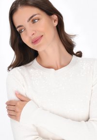 Pulover alb din tricot cu guler rotund. Are paiete strălucitoare pe umeri. Mâneci cu pacient și textură moale.