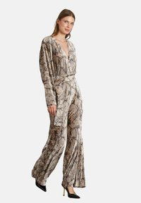 Mono de manga larga en una tela texturizada de tonos tierra, con un escote en V profundo, cintura ceñida y pantalones de pierna ancha. Combinado con tacones negros.