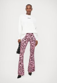 Pantaloni evazați roz cu un model floral negru, asortați cu un pulover alb pe care scrie "STIEGLITZ". Geantă neagră și pantofi cu vârf ascuțit.
