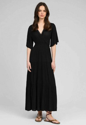 Femme portant une longue robe noire avec des manches aux coudes et un col en V, debout devant un fond uni gris clair, chaussée de sandales marron.