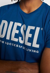 Blå bomullströja med stort vitt "DIESEL"-logotyp och slogan "FOR SUCCESSFULLIVING." Rund halsringning, korta ärmar, slät textur.
